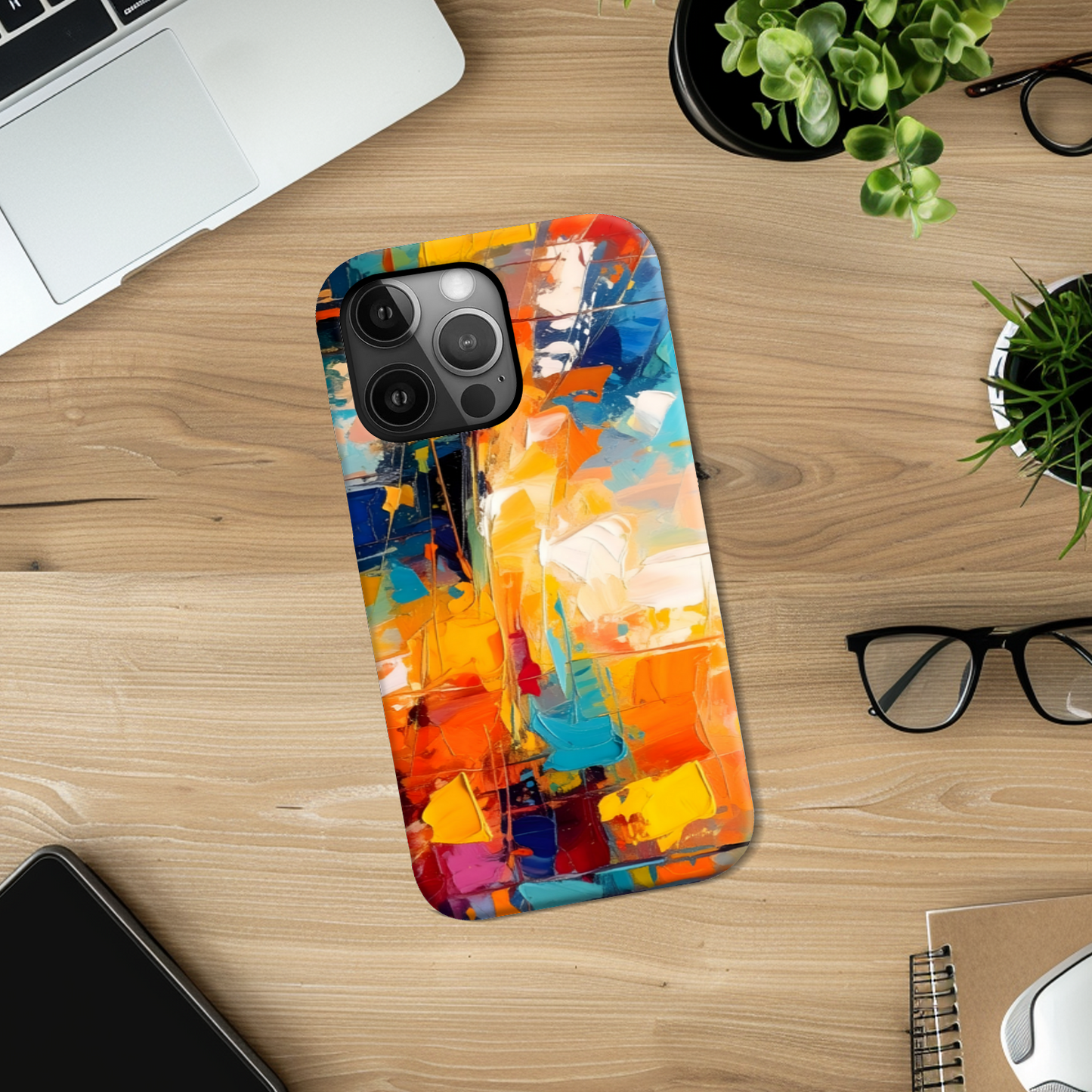 Colorful abstract phone case on a wooden desk with laptop, glasses, and plants.